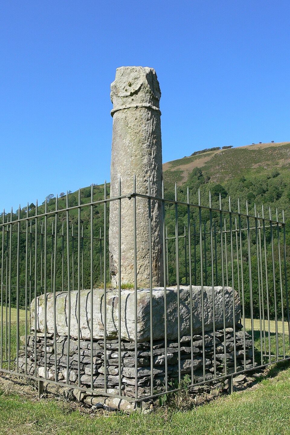 A stone pillar stands on top of a square plinth of grey stones, surrounded by a protective railing. In the background is a verdant, tree-covered hill beneath a bright blue sky.